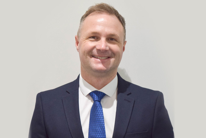 Man (R Harding) in suit in front of white wall smiling at camera